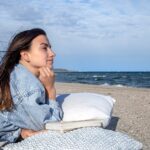 Junge Frau liegt mit Reisekissen am Strand und genießt ihr Outdoorabendteuer